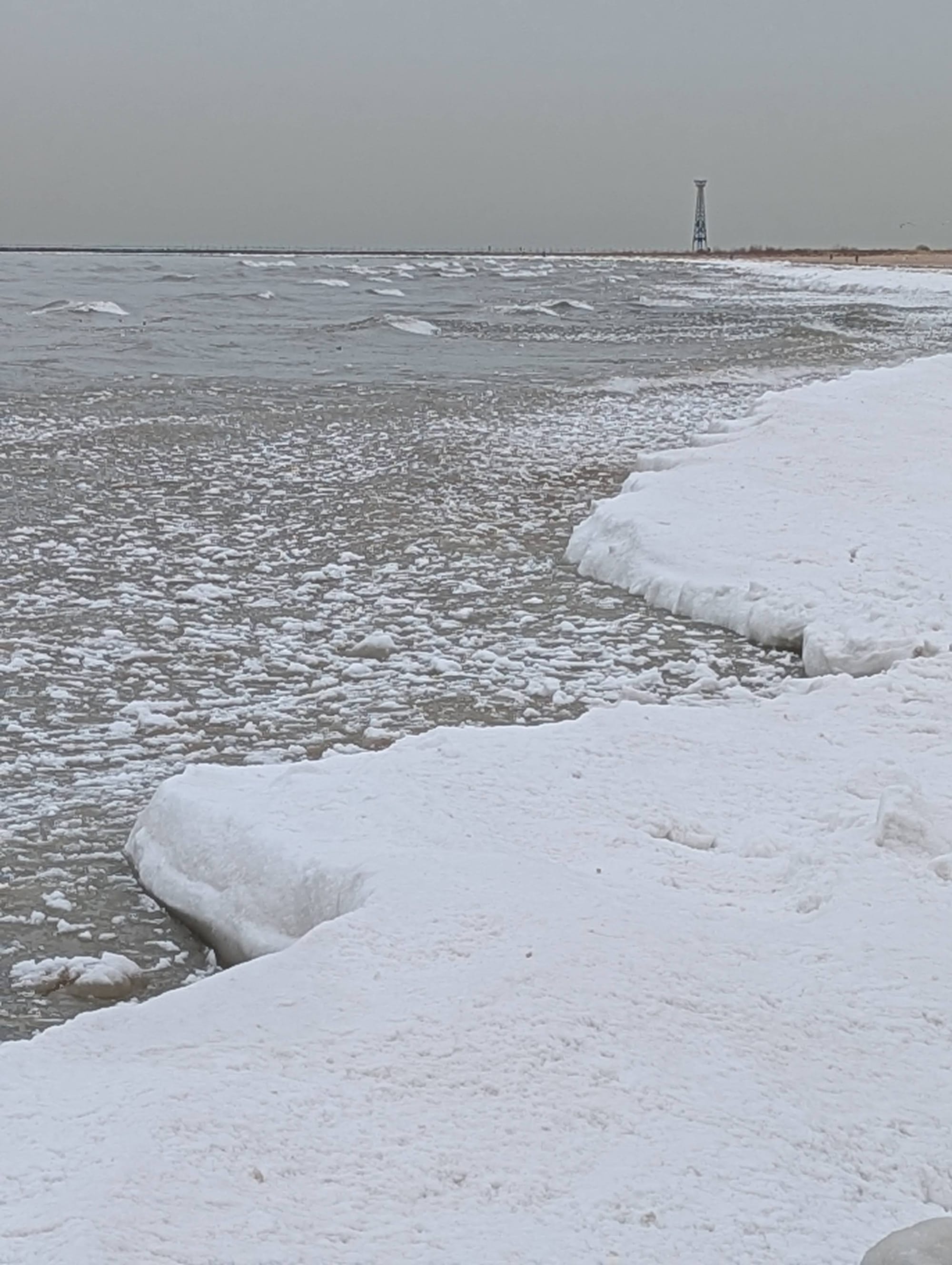 a photo of the shore of Lake Michigan in winter; a zig-zag of snow cuts diagonally across the image and the water is plastered by snowy ice chunks