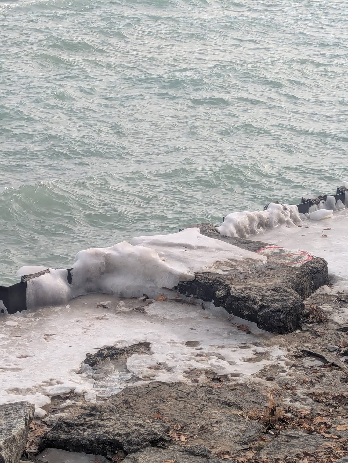 a photo of a teal gray body of water, choppy, cut diagonally by a rocky/concrete barrier at the shore halfway covered in ice