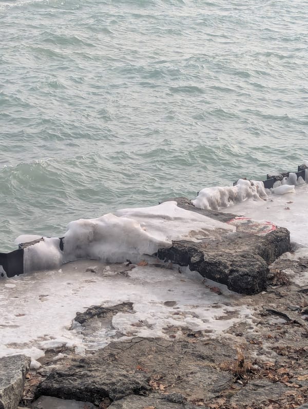 a photo of a teal gray body of water, choppy, cut diagonally by a rocky/concrete barrier at the shore halfway covered in ice